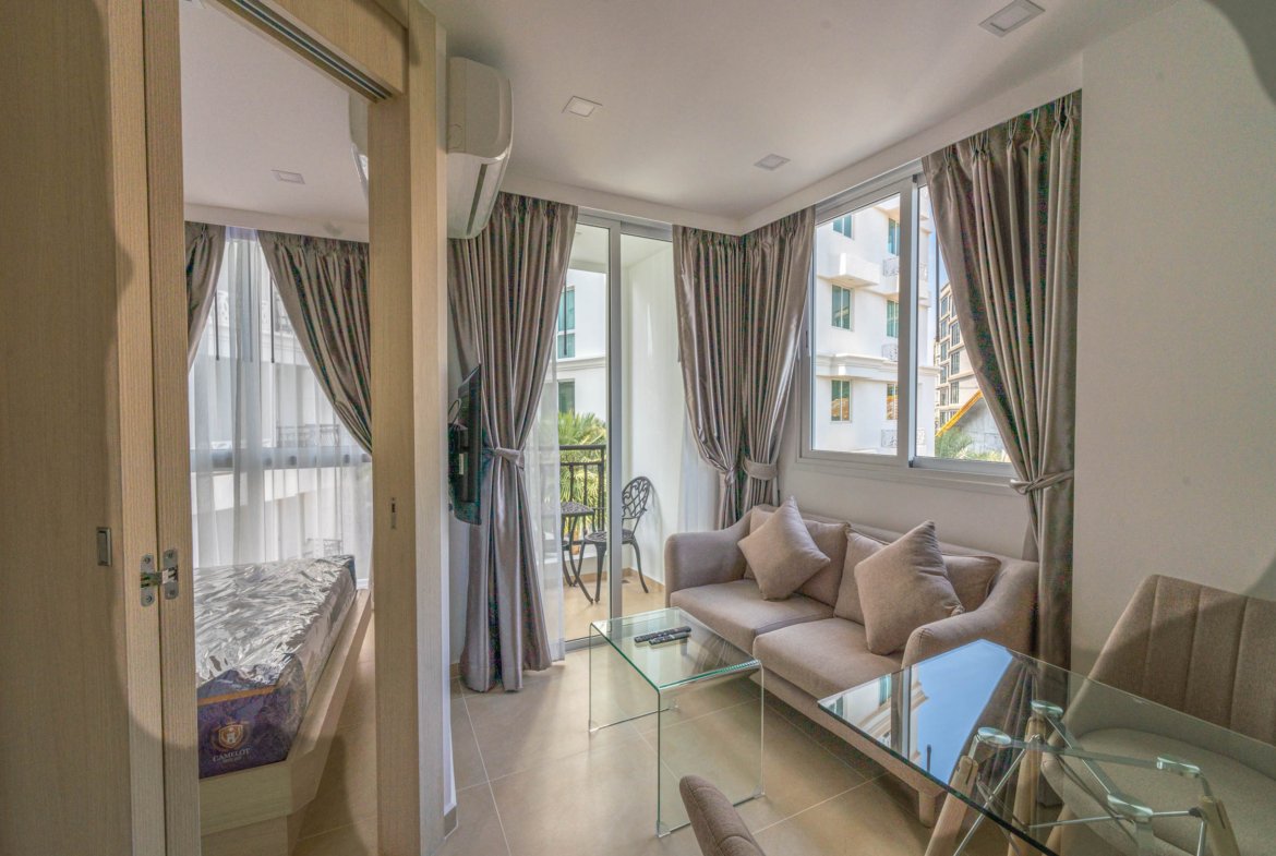 Living room with a glass coffee table, gray sectional sofa, and large windows with gray curtains opening to a balcony seating area on a sunny day
