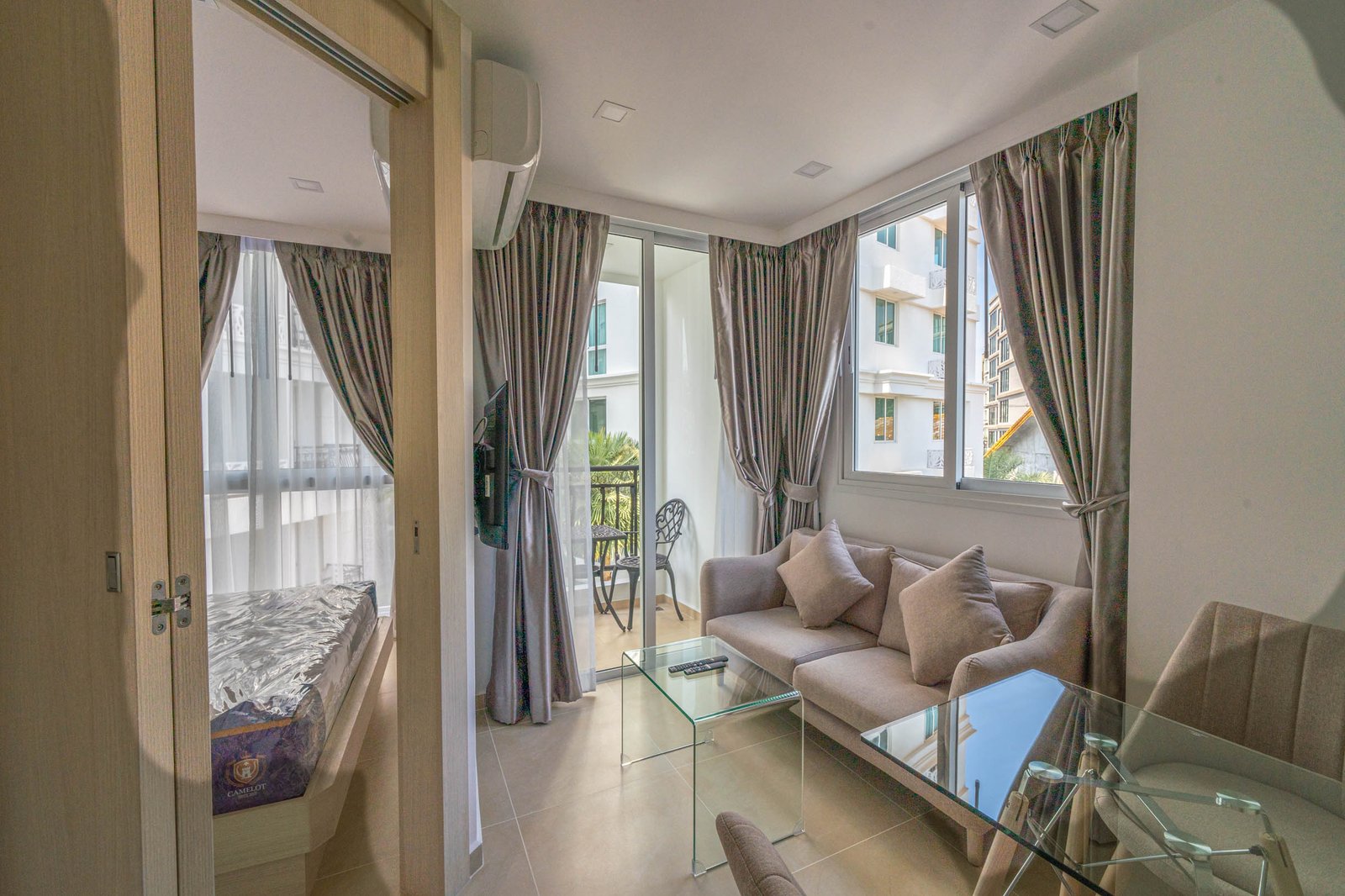Olympus_C306_Rdl79A Living room with a glass coffee table, gray sectional sofa, and large windows with gray curtains opening to a balcony seating area on a sunny day