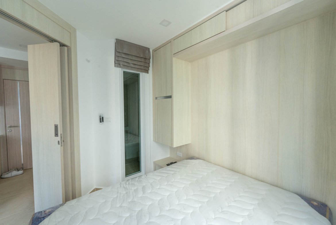 Modern bedroom with light wood built-in wardrobes and a white bedspread; a glass door/window with a gray fabric shade is visible near the wardrobe.