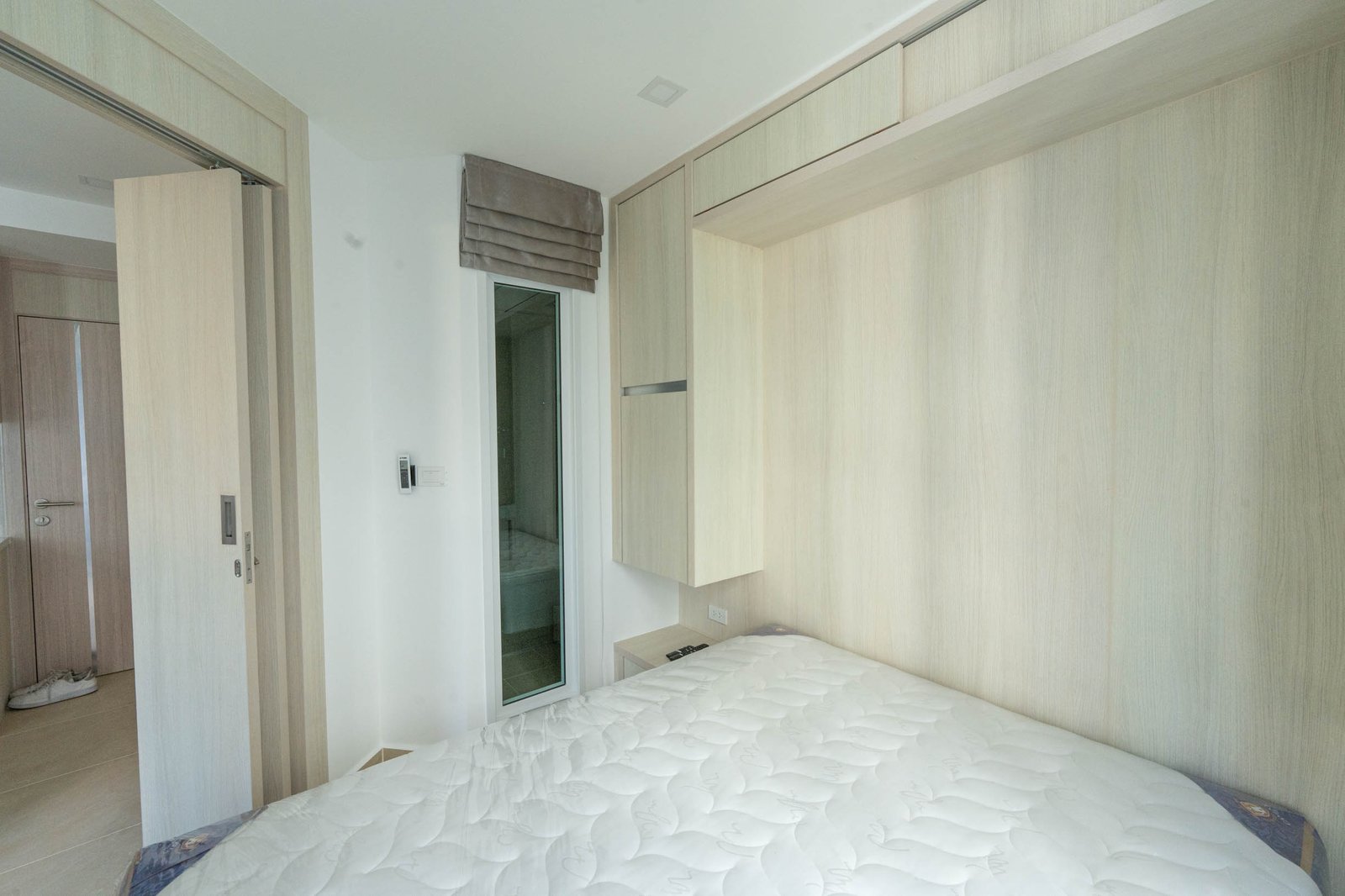 Olympus_C306_Rdl79A_11 Modern bedroom with light wood built-in wardrobes and a white bedspread; a glass door/window with a gray fabric shade is visible near the wardrobe.