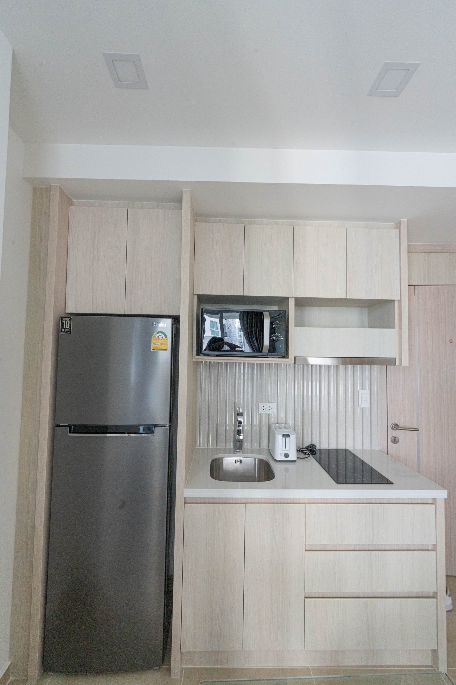 Olympus_C306_Rdl79A_12 Modern kitchen with light wood cabinets, stainless steel fridge, built-in microwave, and white countertop.