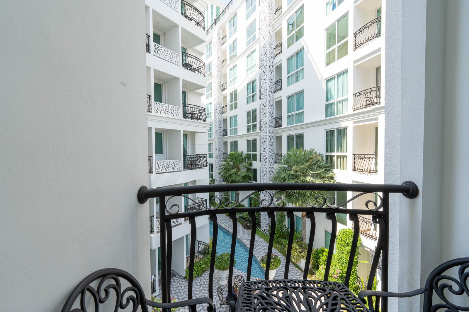 Olympus_C306_Rdl79A_13 View from a balcony overlooking a white modern apartment building with glass windows, ornate black railings, and palm trees in a courtyard.