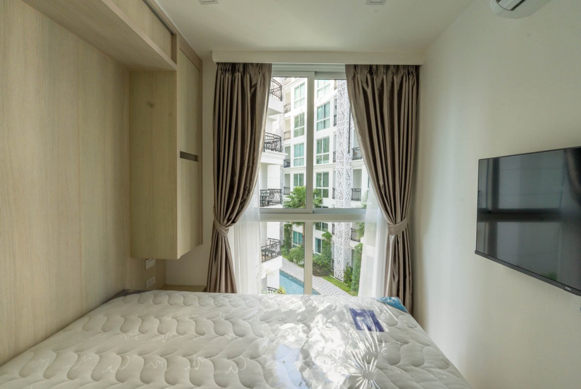 Hotel bedroom interior with a tufted mattress, light wood cabinets on the left, and a large window with brown curtains framing a view of a neighboring building.