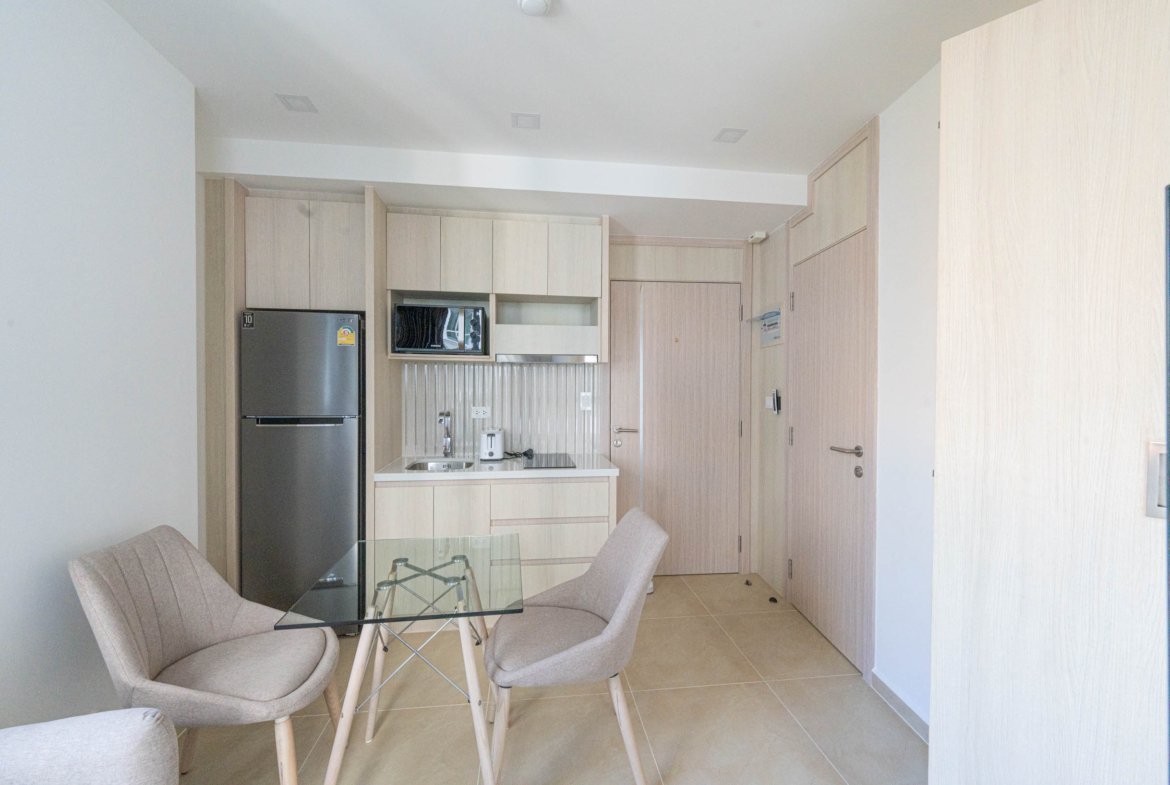 Compact, modern kitchen with light wood cabinets, stainless steel fridge, microwave, and a glass dining table with beige chairs in an open-plan space.