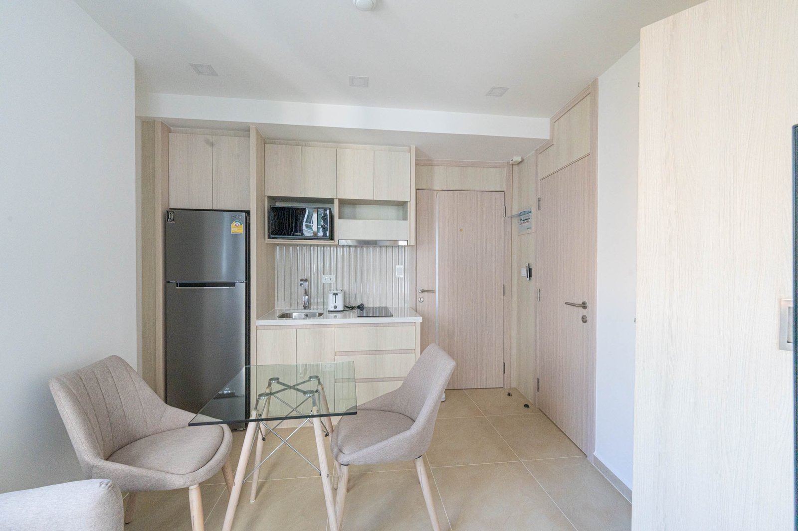Olympus_C306_Rdl79A_3 Compact, modern kitchen with light wood cabinets, stainless steel fridge, microwave, and a glass dining table with beige chairs in an open-plan space.