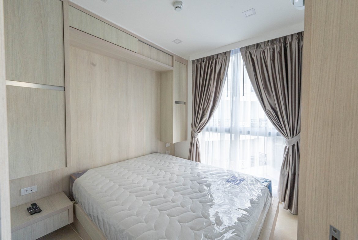 Bedroom with built-in light wood wardrobes along the wall, a mattress on a low bed frame, and a large window with sheer and taupe curtains.