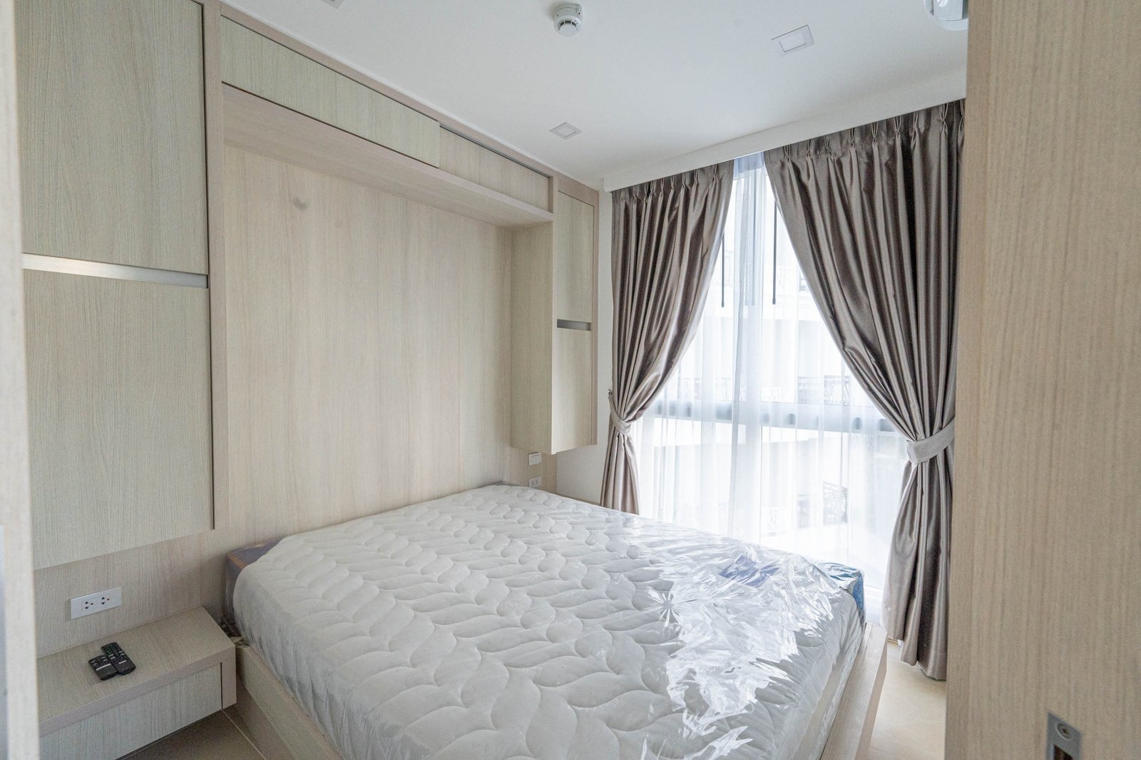 Olympus_C306_Rdl79A_4 Bedroom with built-in light wood wardrobes along the wall, a mattress on a low bed frame, and a large window with sheer and taupe curtains.