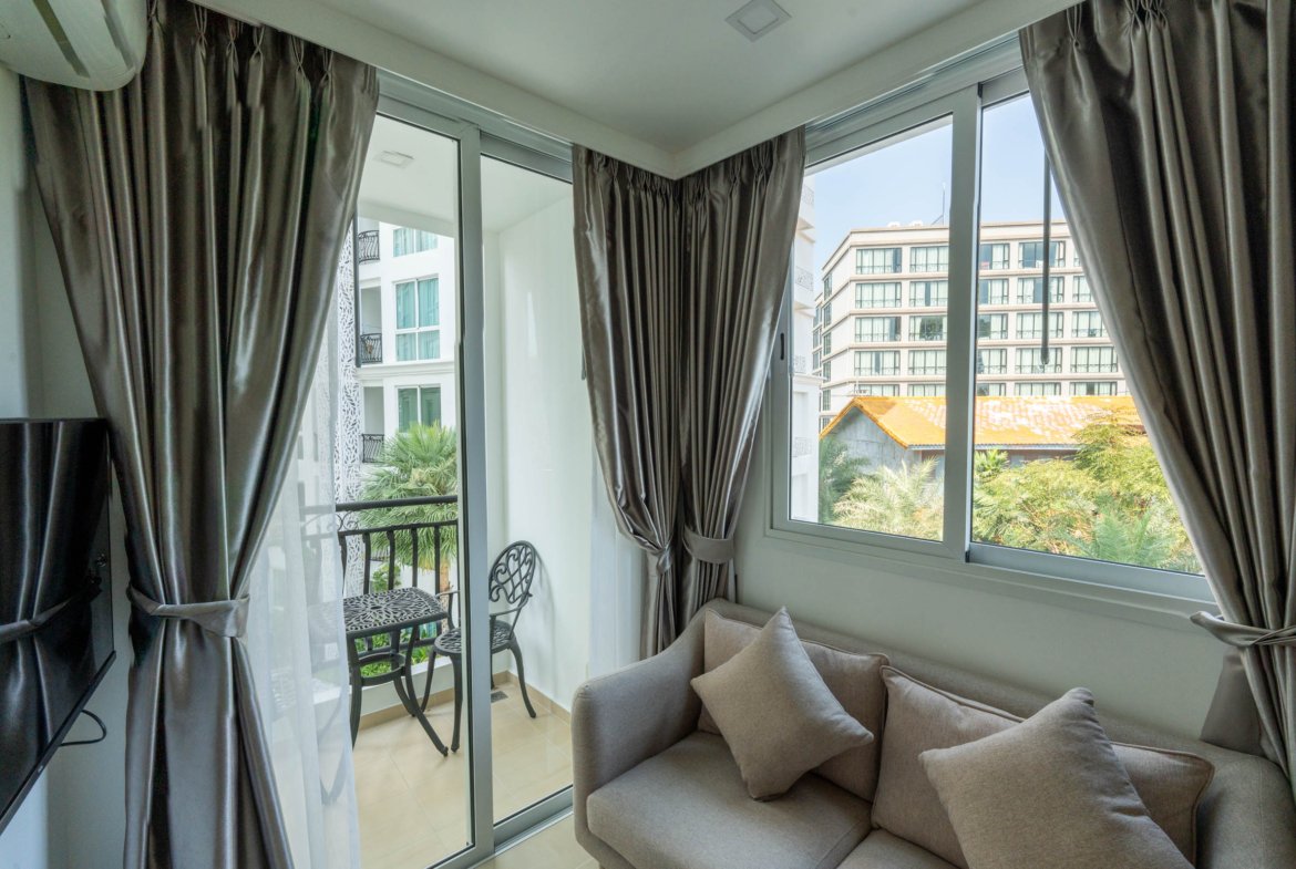 Living room with floor-to-ceiling windows, gray curtains, and a light gray sofa facing a balcony with metal chairs outside.
