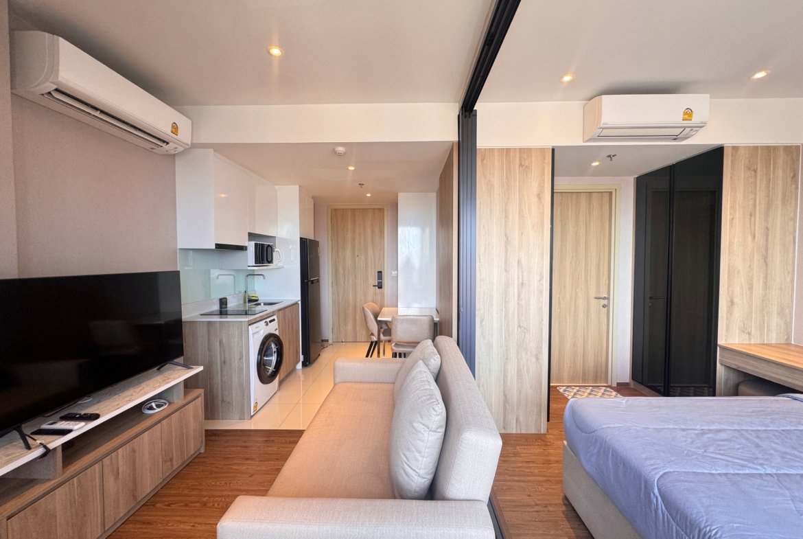 Open-plan living area with a gray sofa facing a TV, kitchenette on the left, and two wall-mounted air conditioners.