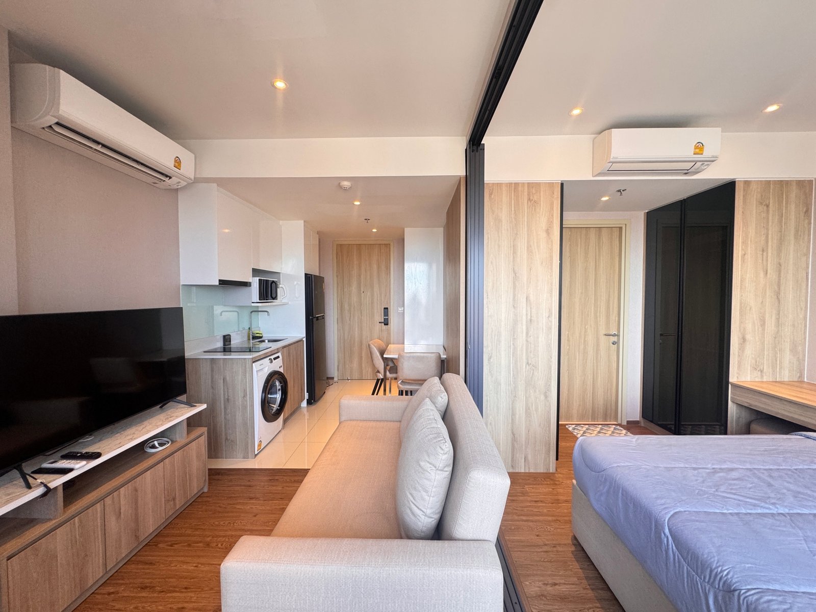 Once_Pattaya_1208_Rent_Rdl295 Open-plan living area with a gray sofa facing a TV, kitchenette on the left, and two wall-mounted air conditioners.