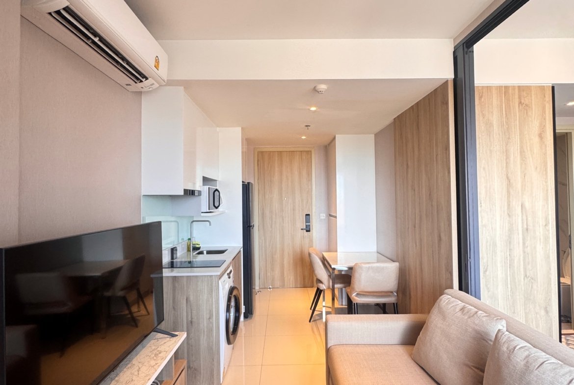 Open-plan living area with a beige sofa facing a wall-mounted TV, and a compact kitchen along the left; AC unit on the wall above.