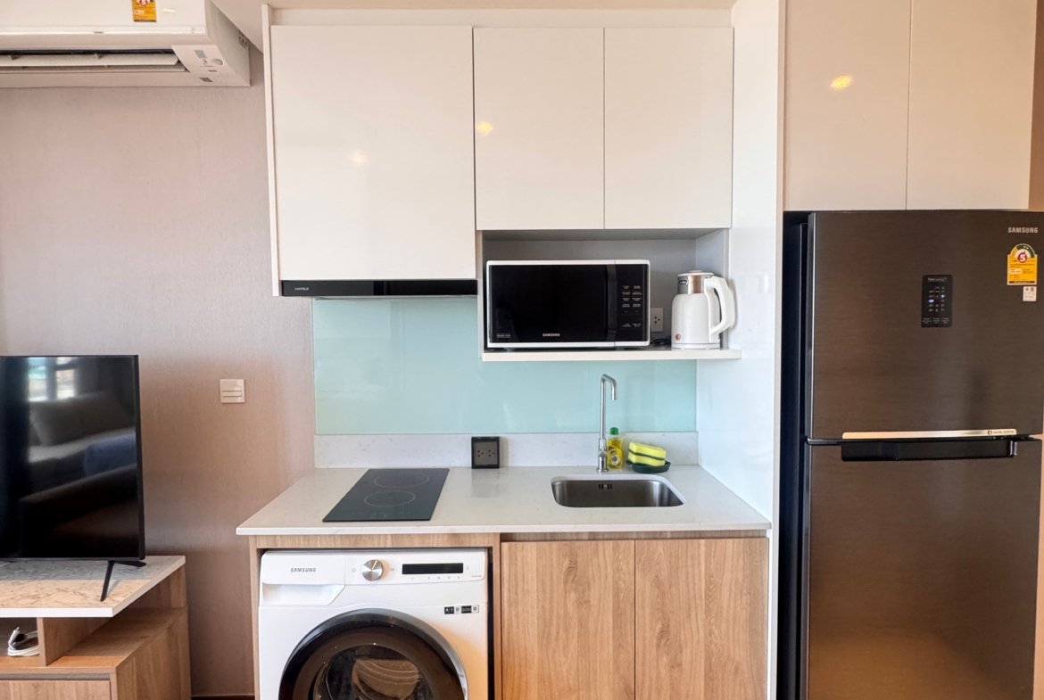 Modern compact kitchen with white upper cabinets, microwave on open shelf, kettle, and a stainless fridge to the right