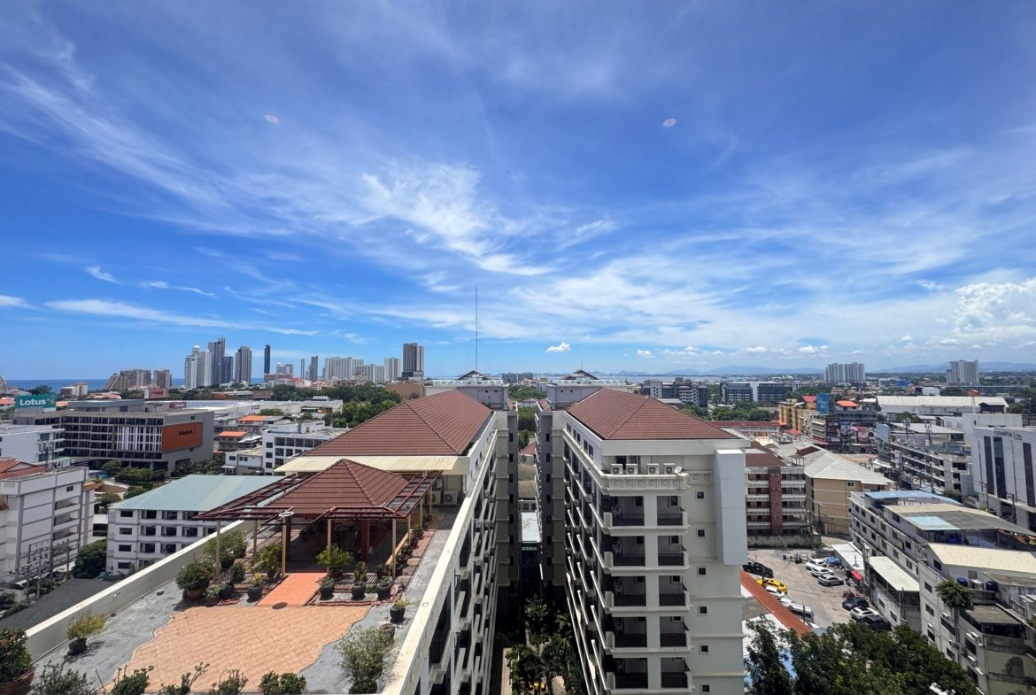City skyline under a bright blue sky with wispy clouds; foreground shows rooftops and a multi-story residential building terrace.