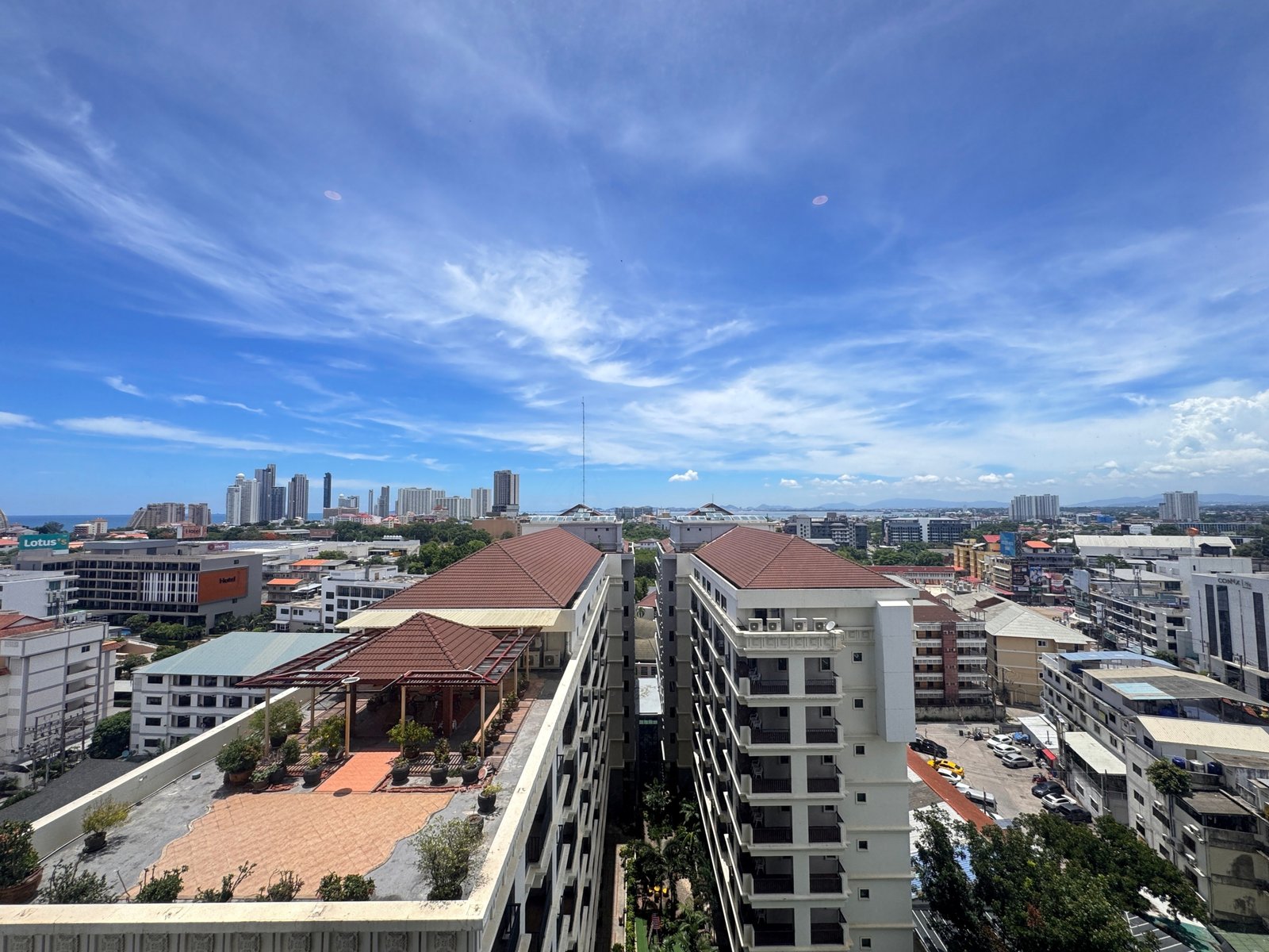 Once_Pattaya_1208_Rent_Rdl295_18 City skyline under a bright blue sky with wispy clouds; foreground shows rooftops and a multi-story residential building terrace.