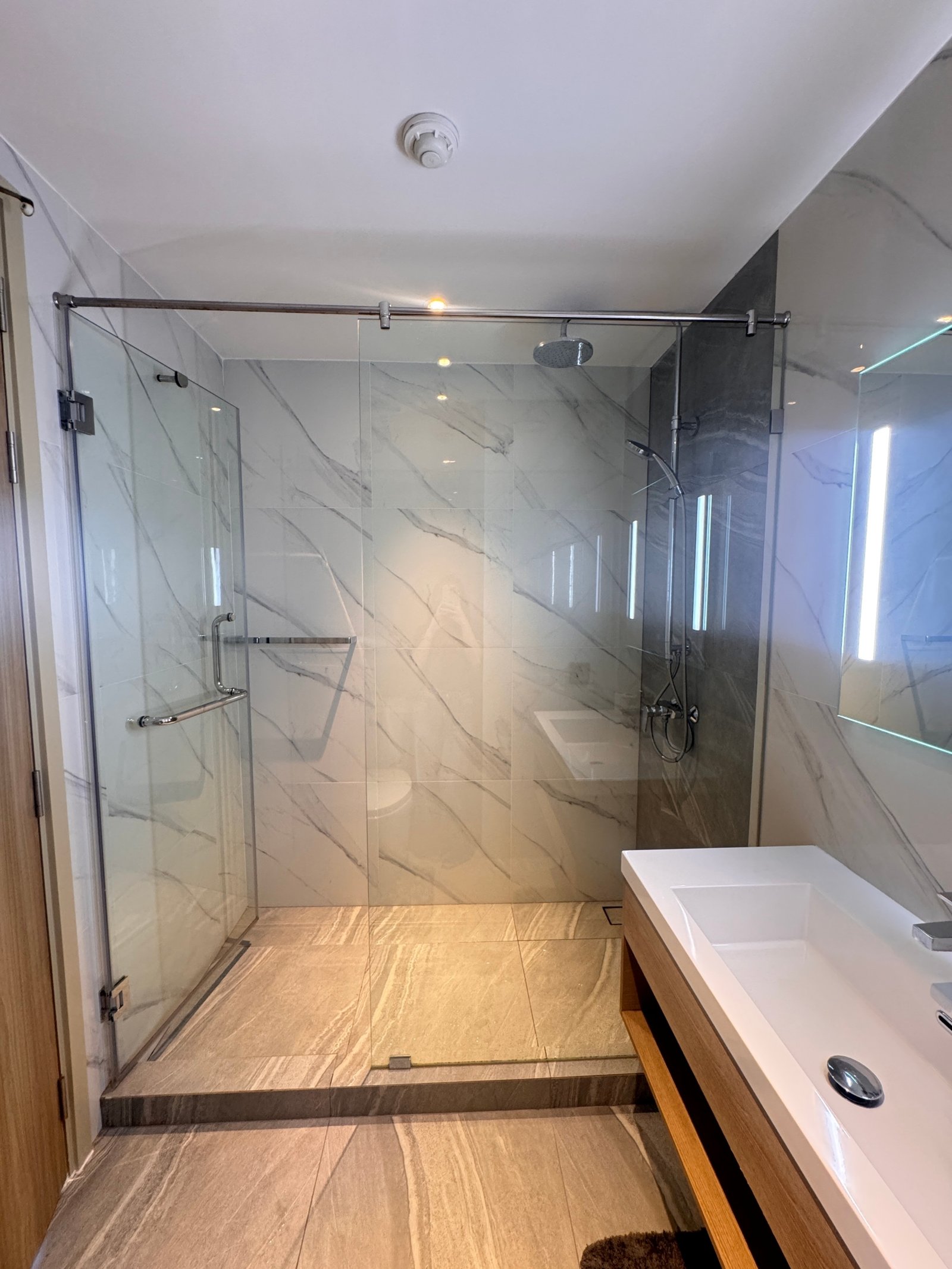 Once_Pattaya_1208_Rent_Rdl295_6 Modern bathroom featuring a glass shower enclosure with marble walls and a rainfall showerhead, plus a white rectangular sink and wooden vanity.