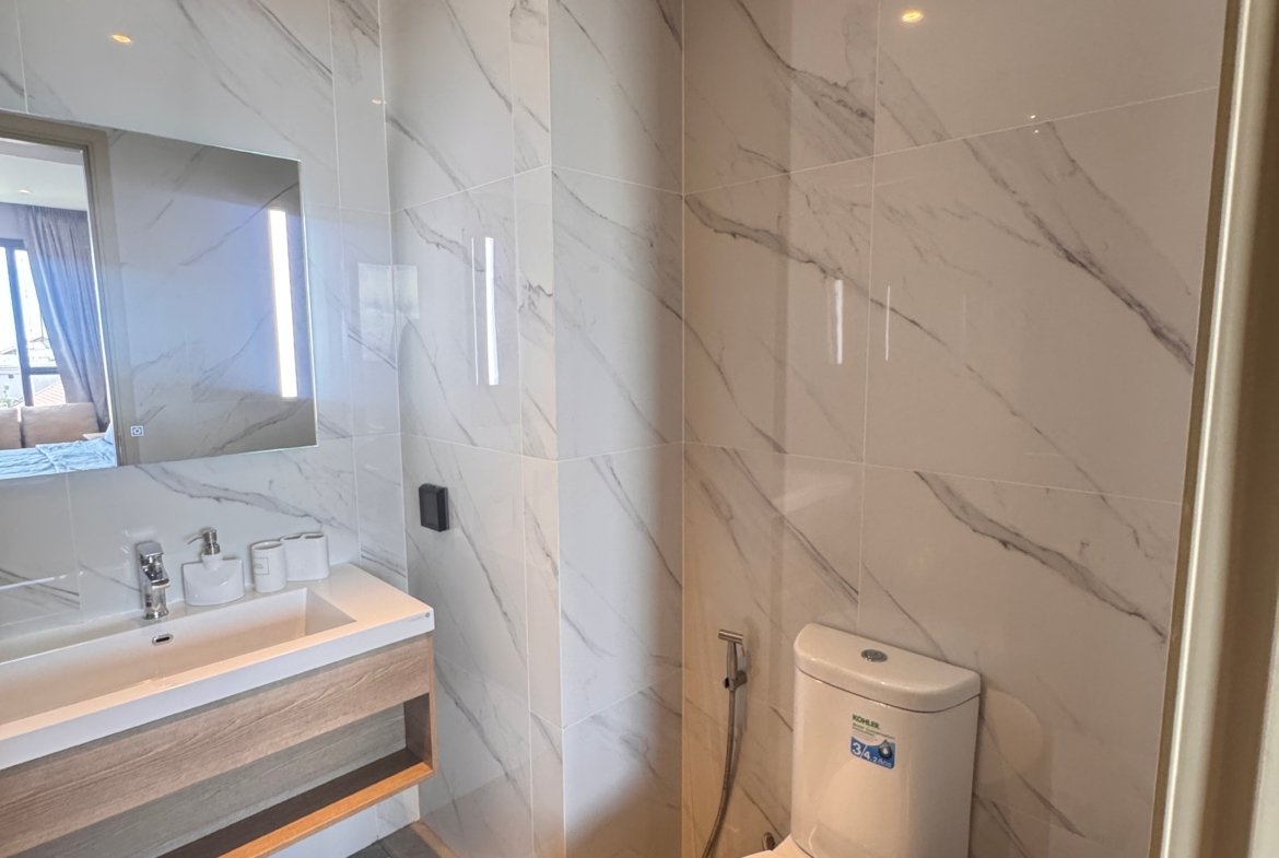 Modern bathroom with marble tiled walls, wall-mounted sink, large mirror, and toilet with handheld bidet sprayer