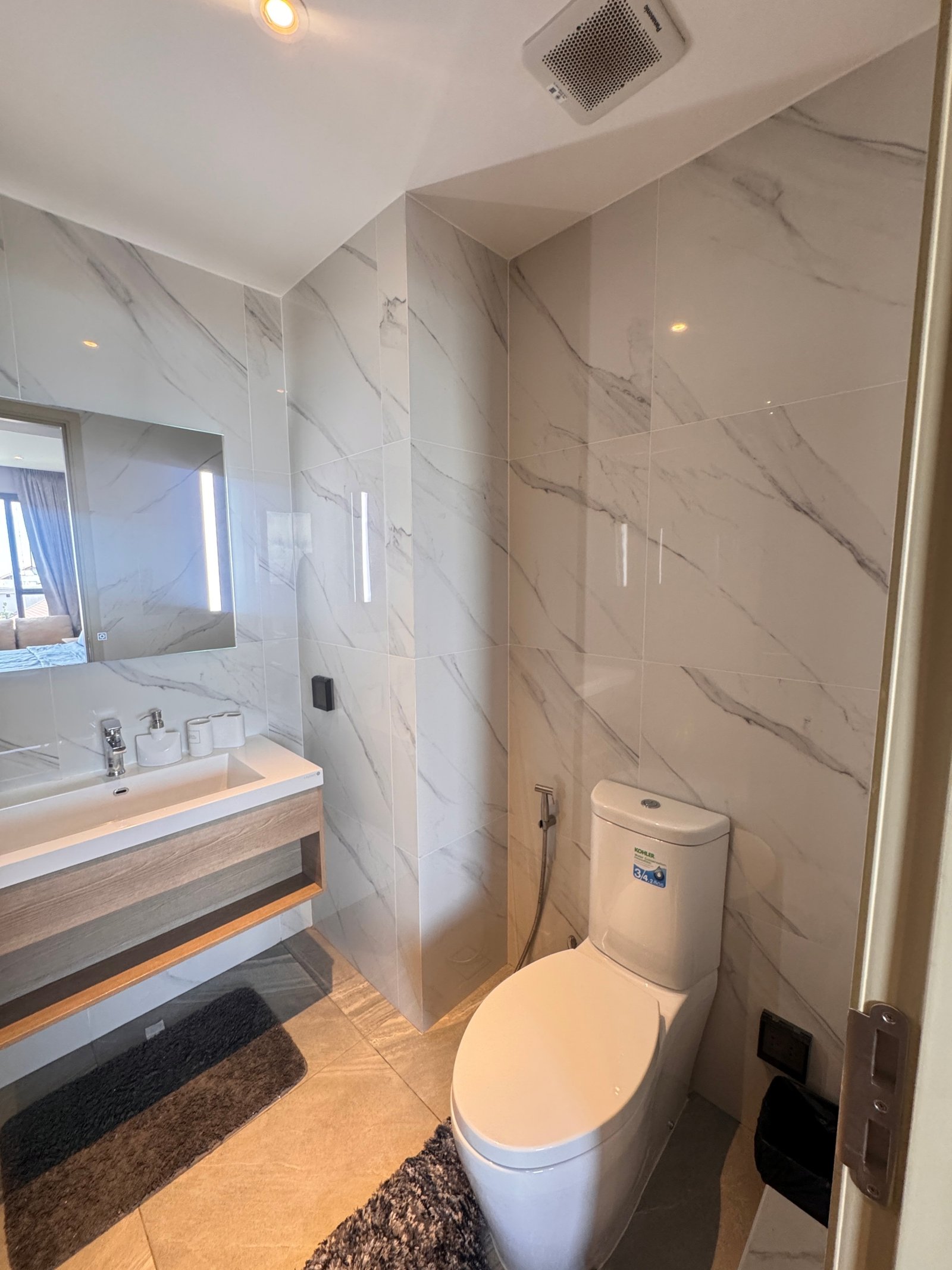 Once_Pattaya_1208_Rent_Rdl295_7 Modern bathroom with marble tiled walls, wall-mounted sink, large mirror, and toilet with handheld bidet sprayer