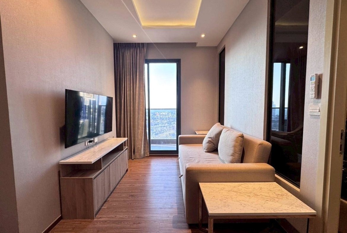 Modern hotel living room with a beige sofa, wall-mounted TV, marble coffee table, and a balcony door showing a city view.