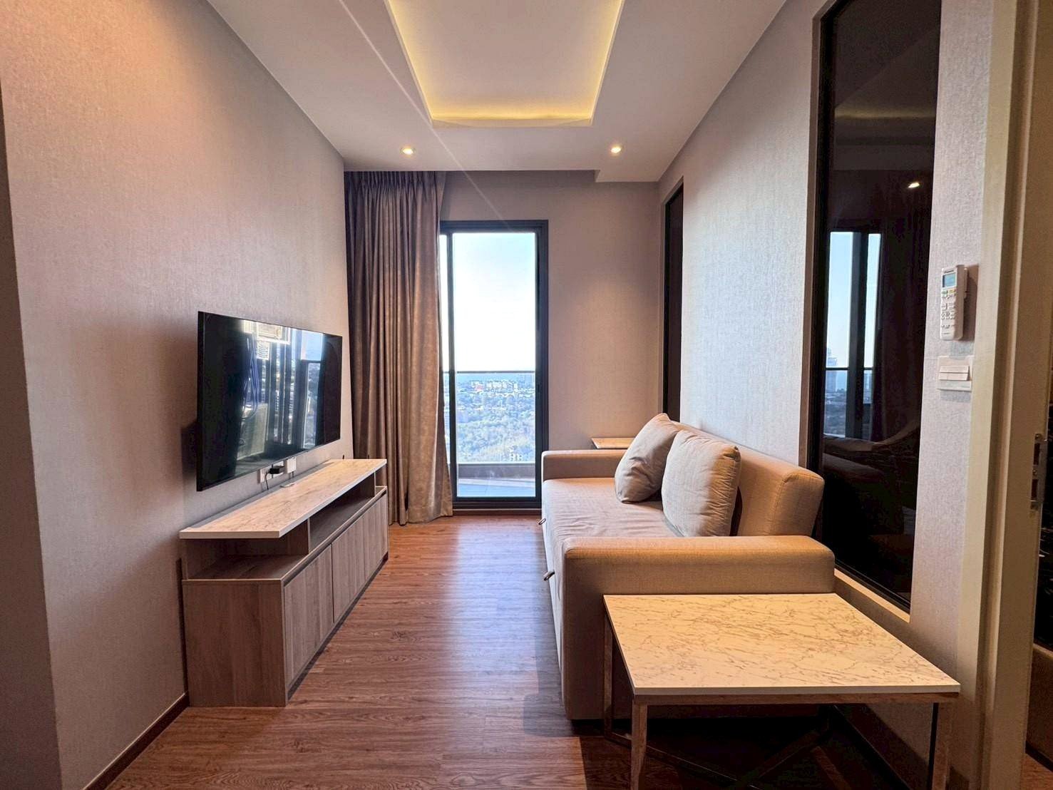 Modern hotel living room with a beige sofa, wall-mounted TV, marble coffee table, and a balcony door showing a city view.
