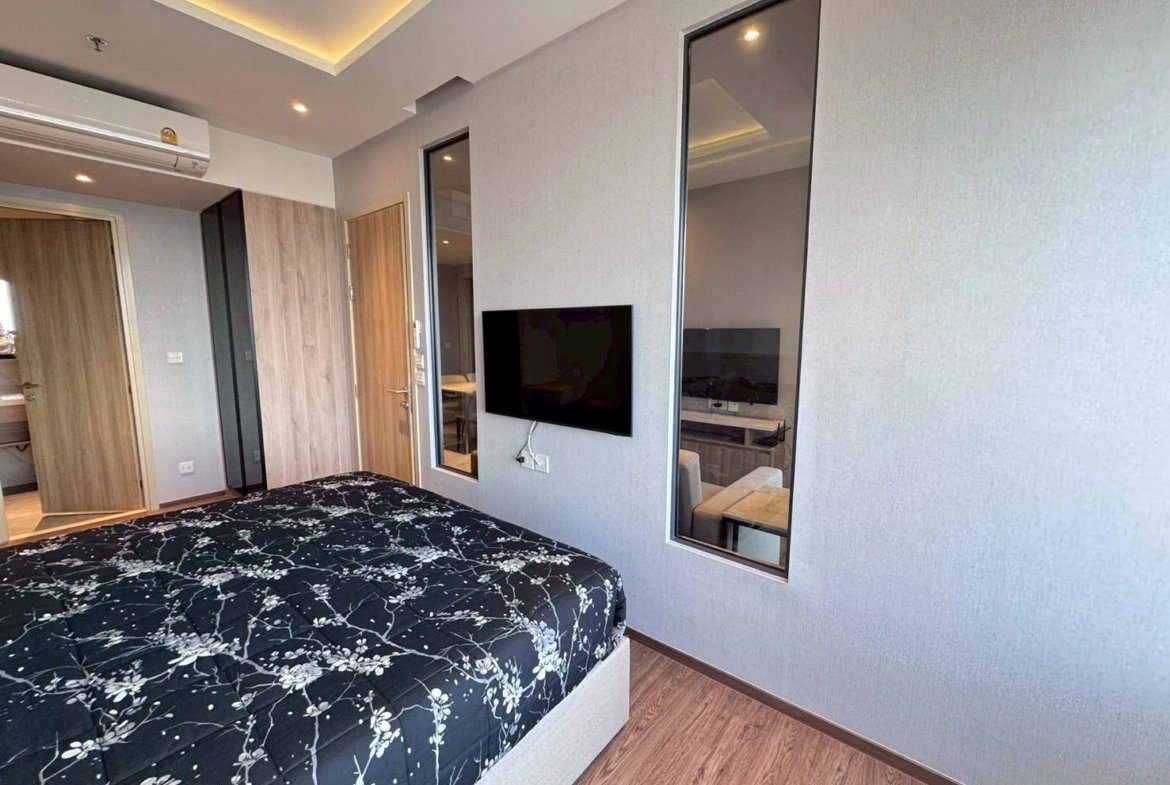Bedroom in a modern hotel: queen bed with black floral bedding, wood floor, wall-mounted TV, and tall windows.