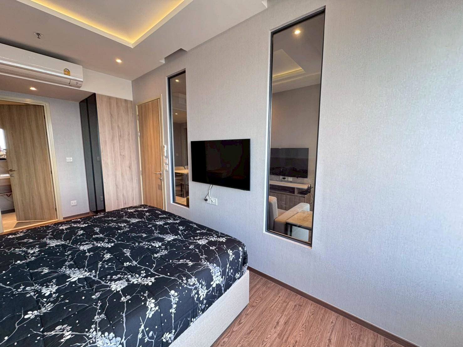 Bedroom in a modern hotel: queen bed with black floral bedding, wood floor, wall-mounted TV, and tall windows.