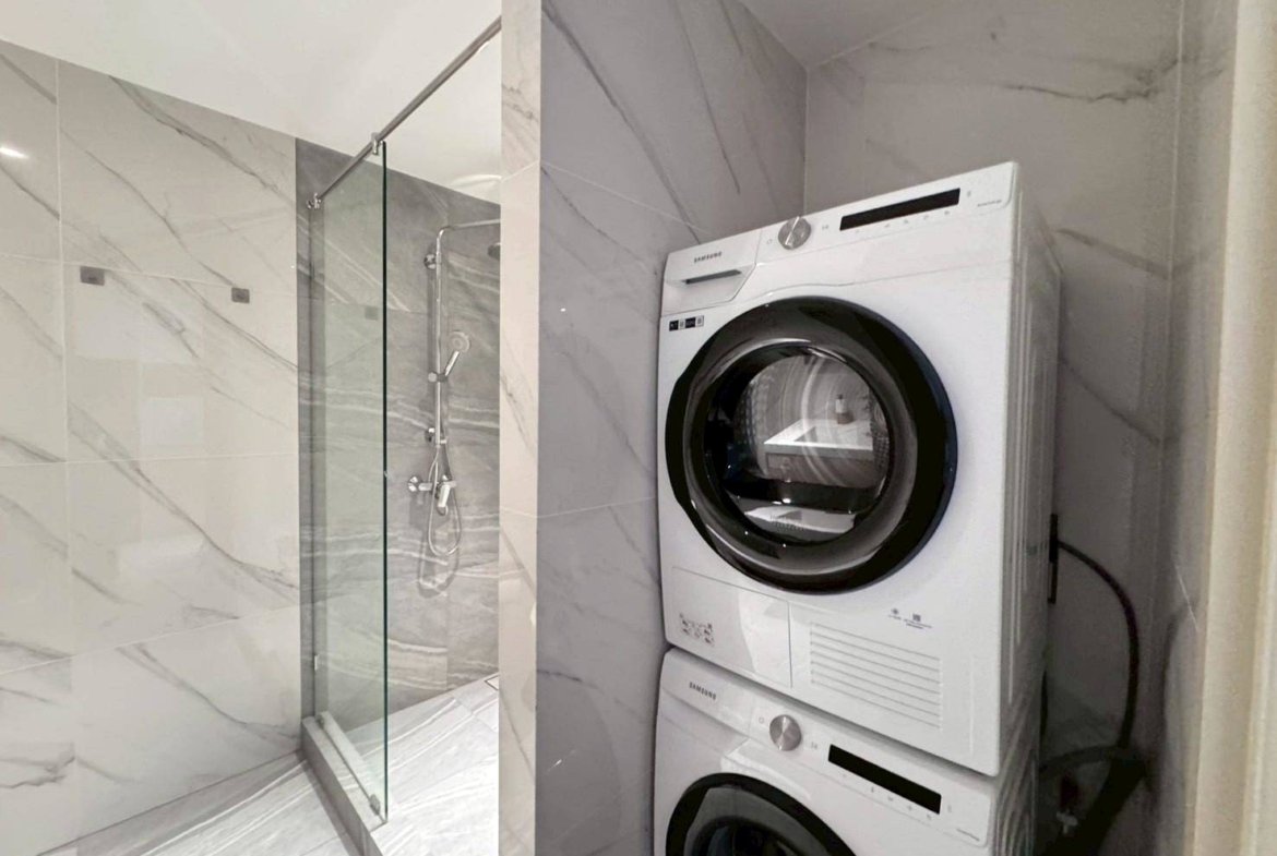 Stacked white washing machine and dryer beside a glass-enclosed marble-tiled shower in a modern bathroom.