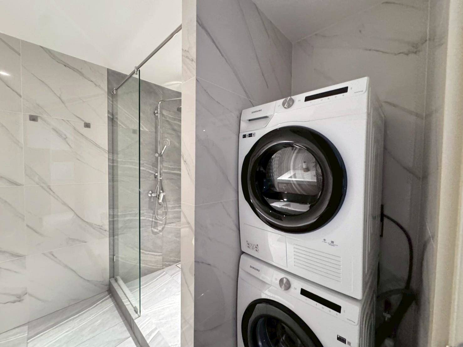Stacked white washing machine and dryer beside a glass-enclosed marble-tiled shower in a modern bathroom.