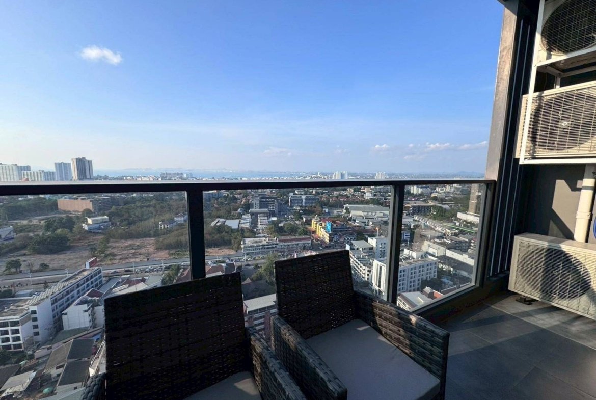 Balcony with wicker chairs overlooking a cityscape of low and mid-rise buildings under a clear blue sky.