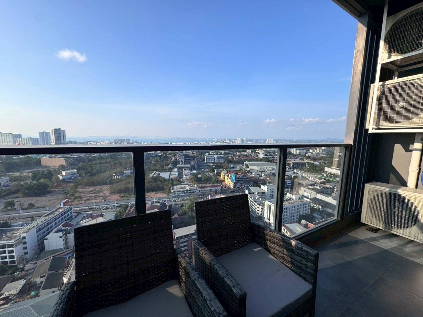 Balcony with wicker chairs overlooking a cityscape of low and mid-rise buildings under a clear blue sky.