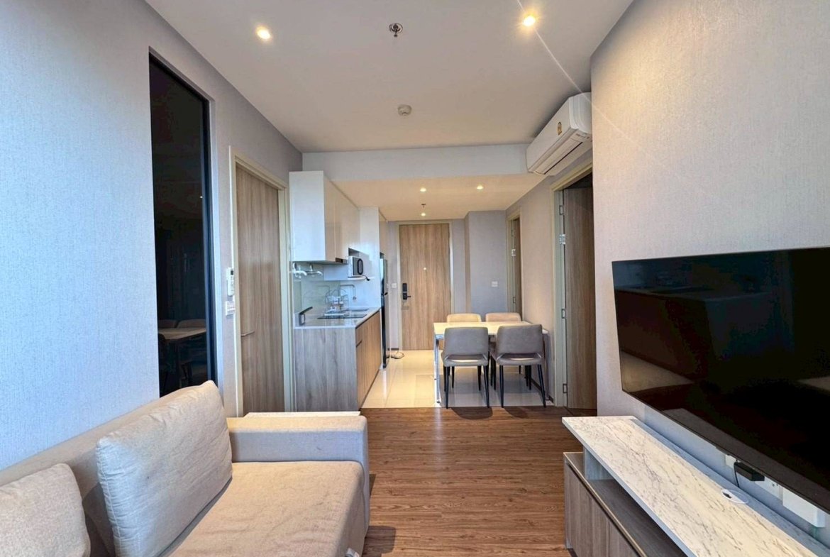 Open-concept living area with sofa, wall-mounted TV, and marble TV stand facing a kitchen and dining nook in a modern apartment.