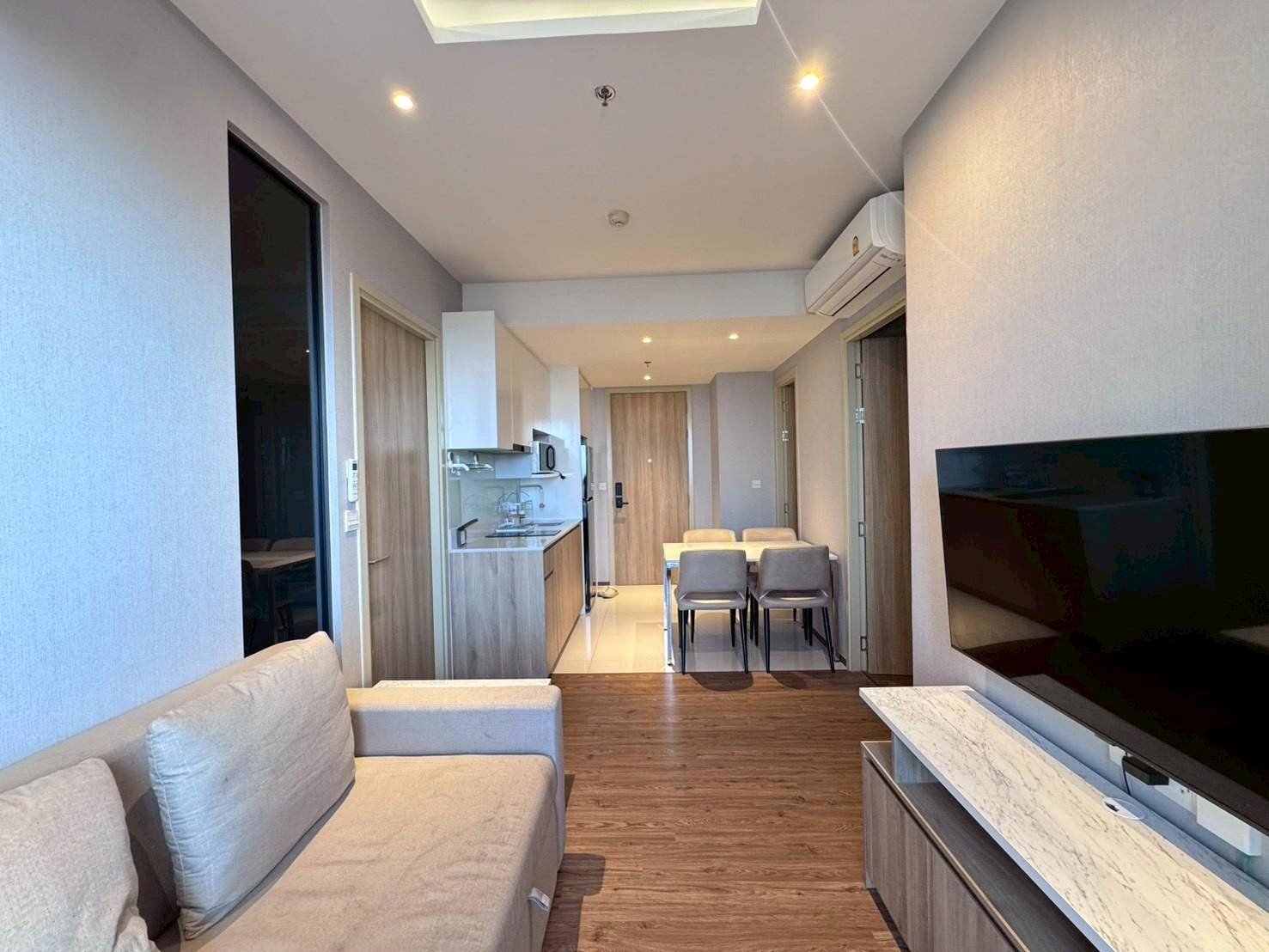 Open-concept living area with sofa, wall-mounted TV, and marble TV stand facing a kitchen and dining nook in a modern apartment.