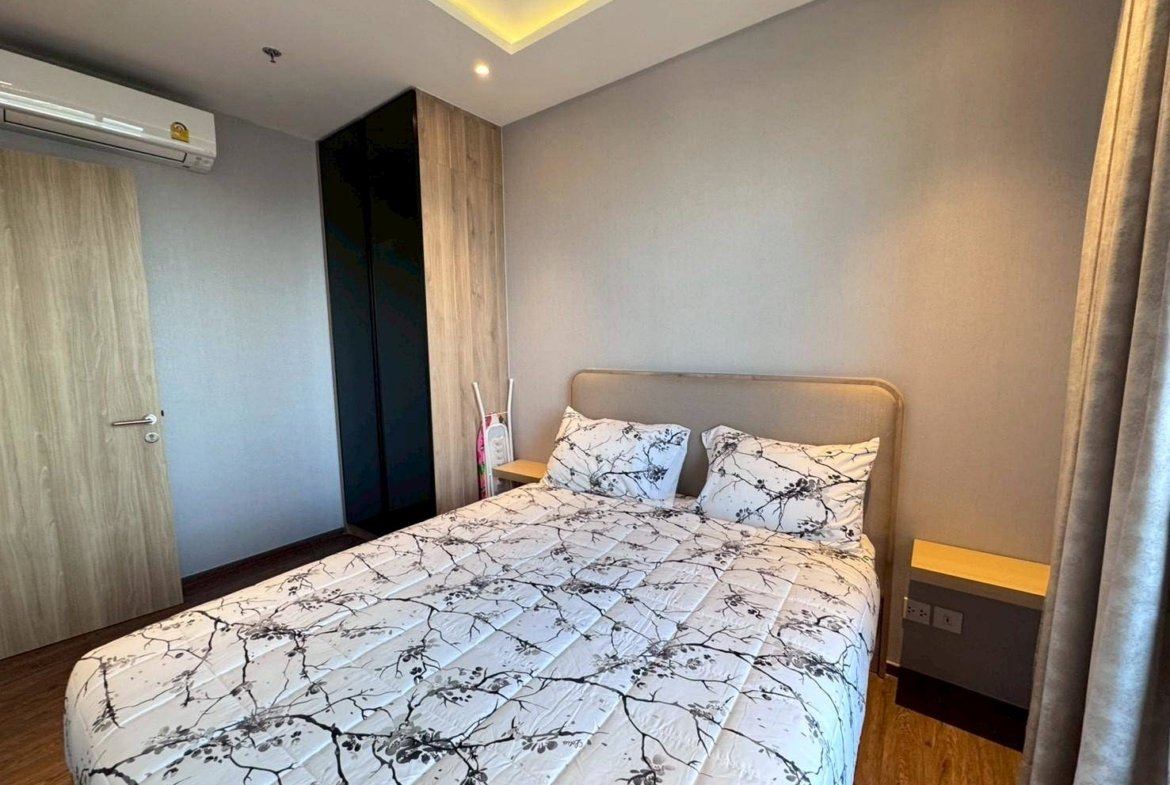 Small modern bedroom with a double bed, gray walls, and a wooden wardrobe along the left wall. Nearby are two nightstands and a window with beige curtains.