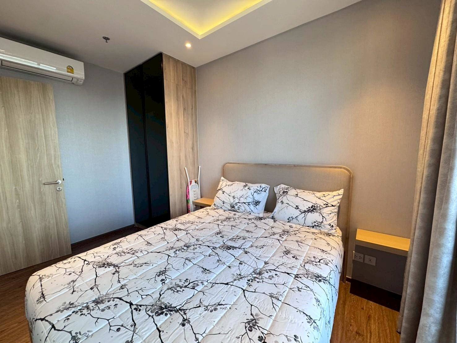 Small modern bedroom with a double bed, gray walls, and a wooden wardrobe along the left wall. Nearby are two nightstands and a window with beige curtains.