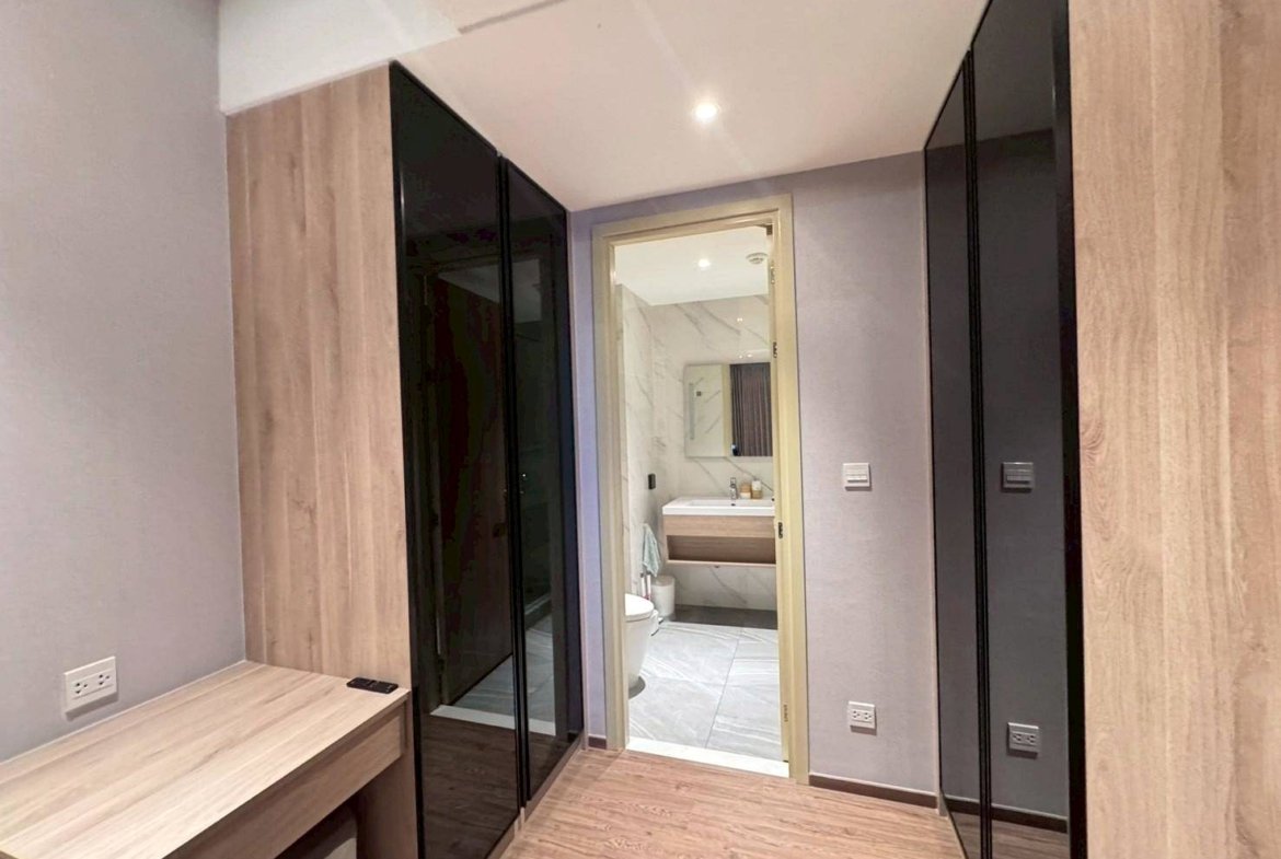 Narrow dressing area with light wood flooring and built‑in wardrobes with black glass doors, desk on the left, and a doorway to a marble-tiled bathroom.