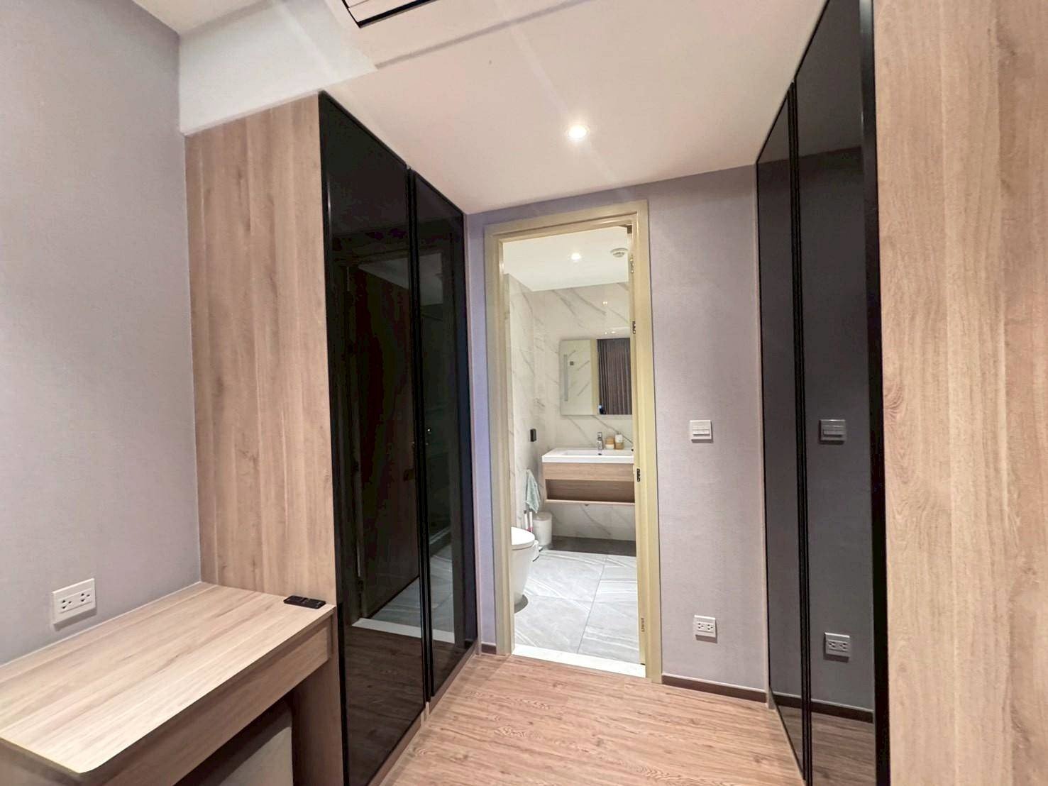 Narrow dressing area with light wood flooring and built‑in wardrobes with black glass doors, desk on the left, and a doorway to a marble-tiled bathroom.