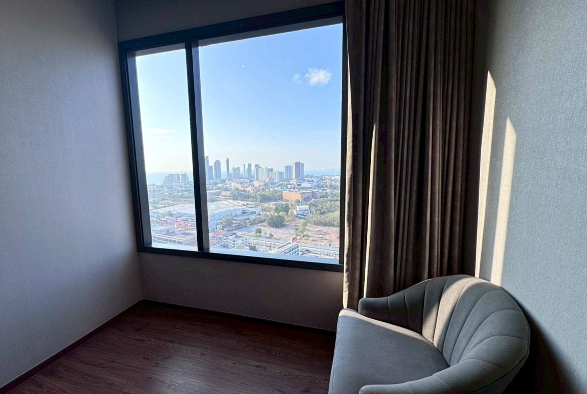 Corner room with a large window showing a city skyline; dark curtains on the right and a gray curved chair in the foreground.
