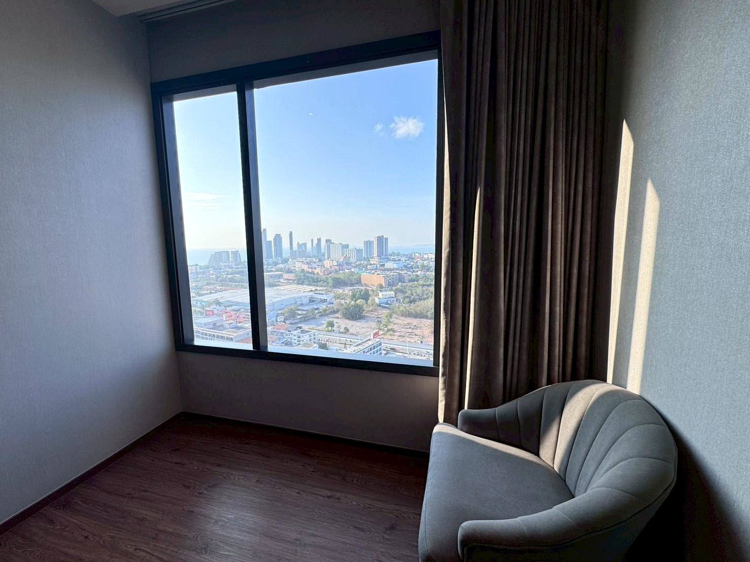 Corner room with a large window showing a city skyline; dark curtains on the right and a gray curved chair in the foreground.