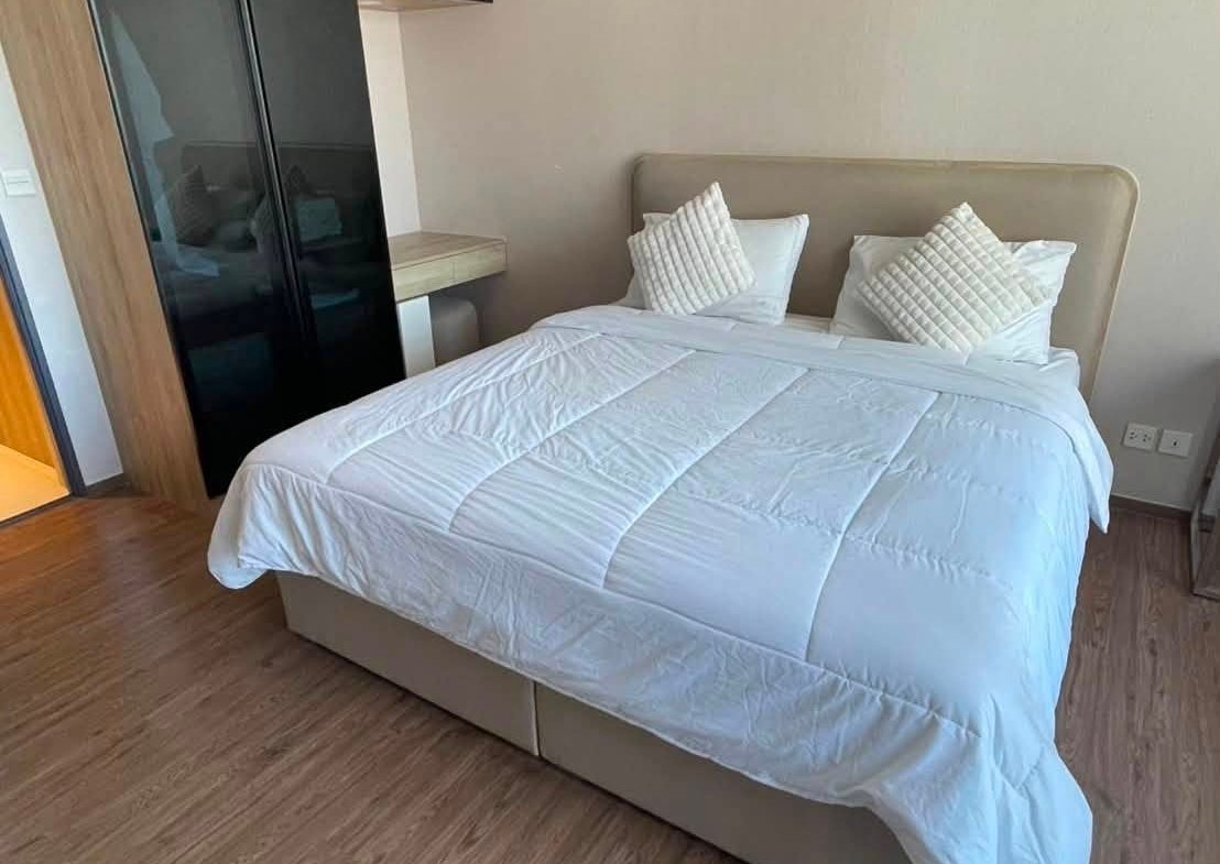 Double bed with white comforter and two textured pillows in a modern bedroom, beige headboard and wooden floor, wardrobe on the left.