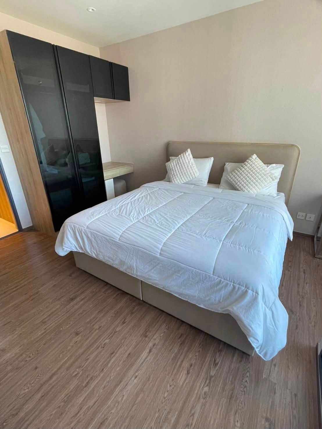 Double bed with white comforter and two textured pillows in a modern bedroom, beige headboard and wooden floor, wardrobe on the left.