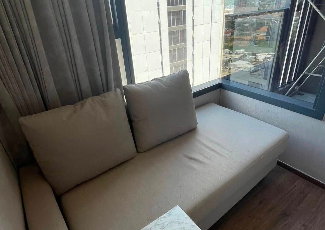 Beige sectional sofa with two pillows beside a marble-topped table, by a large window letting in daylight.