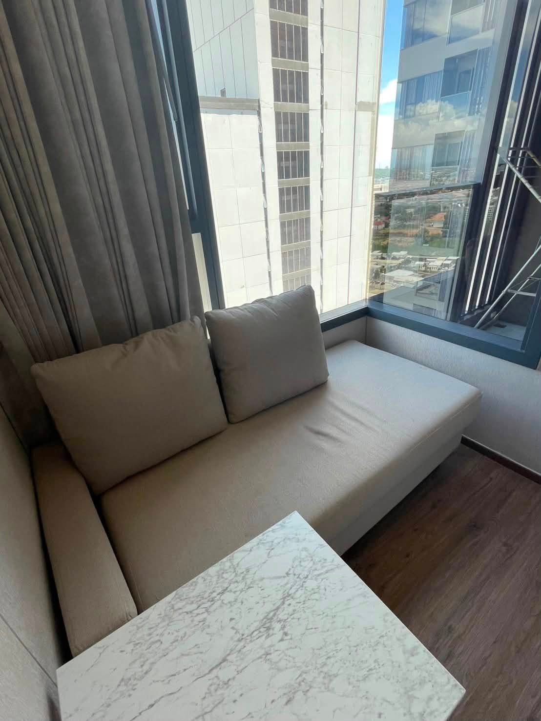 Beige sectional sofa with two pillows beside a marble-topped table, by a large window letting in daylight.