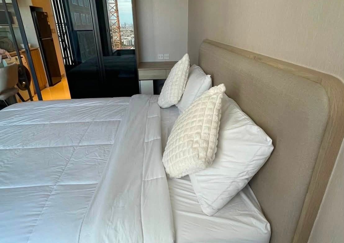 Bedroom with white bedding and two textured pillows, a beige headboard, a black glossy wardrobe, a small desk, and a city view through the window.