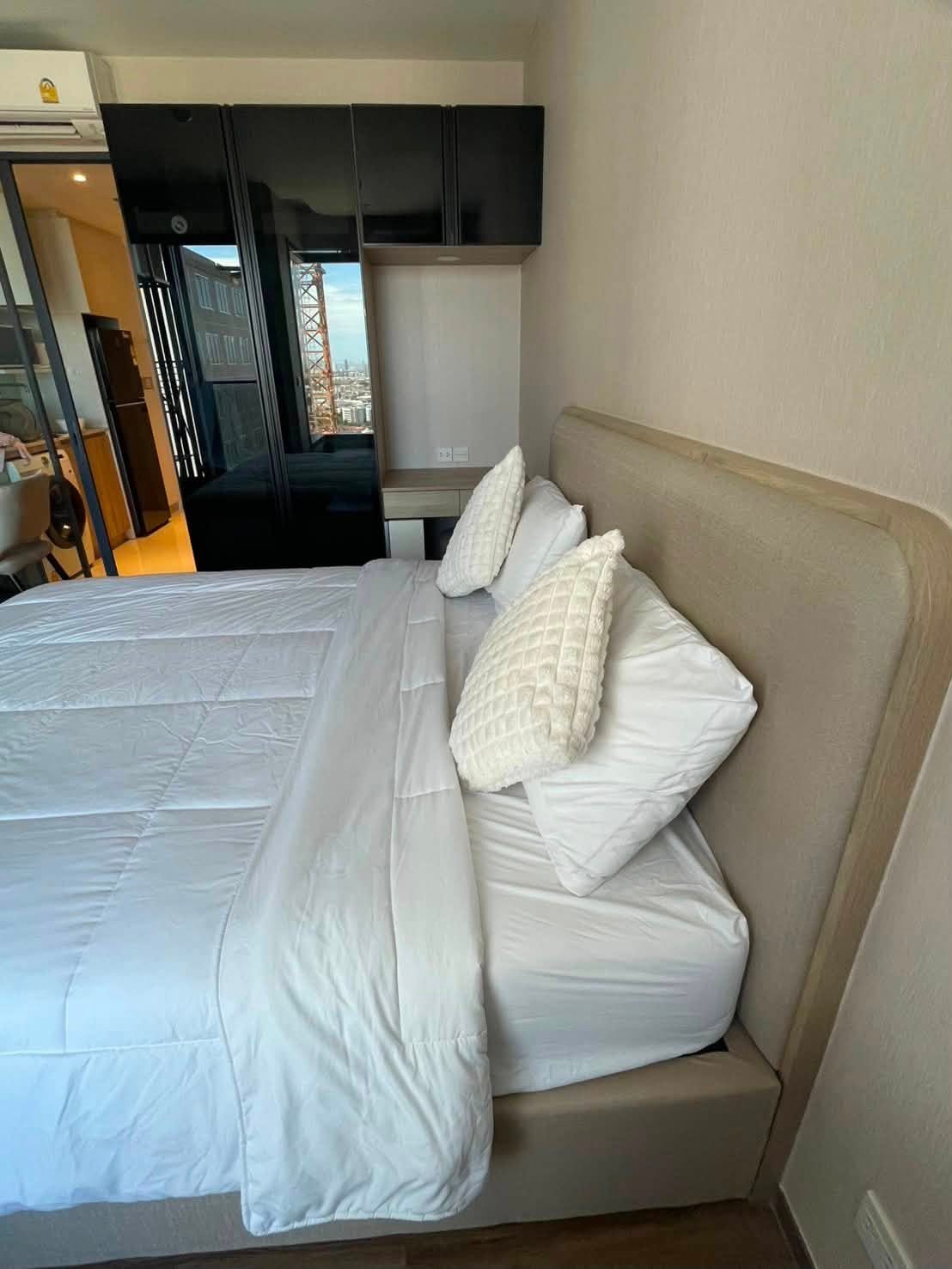 Bedroom with white bedding and two textured pillows, a beige headboard, a black glossy wardrobe, a small desk, and a city view through the window.