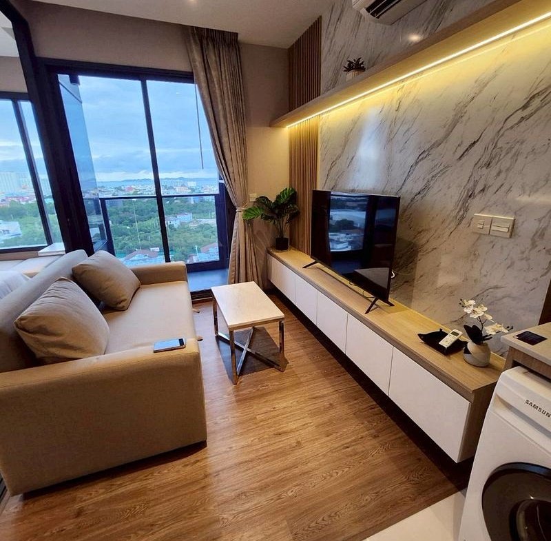 Cozy modern living room with a beige sofa facing a flat-screen TV, marble-pattern wall, and floor-to-ceiling windows revealing a city view.