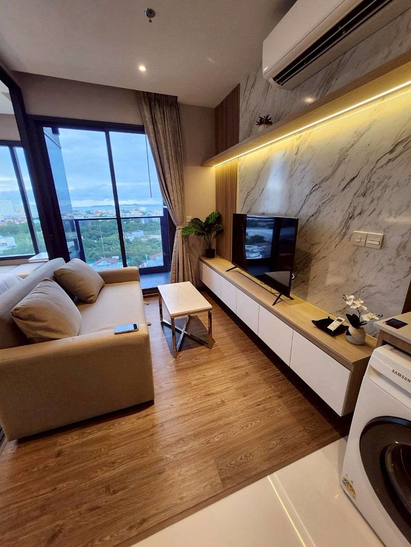 Cozy modern living room with a beige sofa facing a flat-screen TV, marble-pattern wall, and floor-to-ceiling windows revealing a city view.