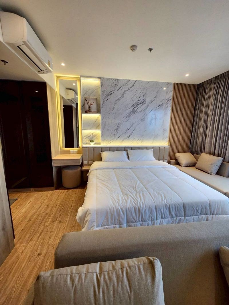 Modern hotel room with a large bed in white bedding, a marble accent wall, and a gray sofa seating area beside it.