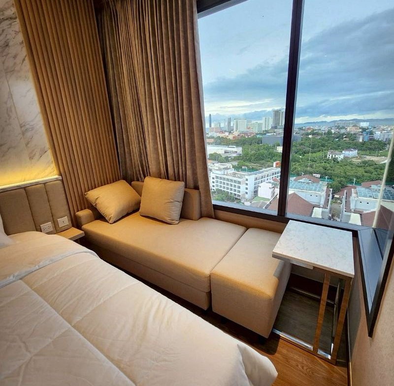 Hotel room corner with a beige sofa bed by a large window overlooking a cityscape. Fixed curtains frame the view.