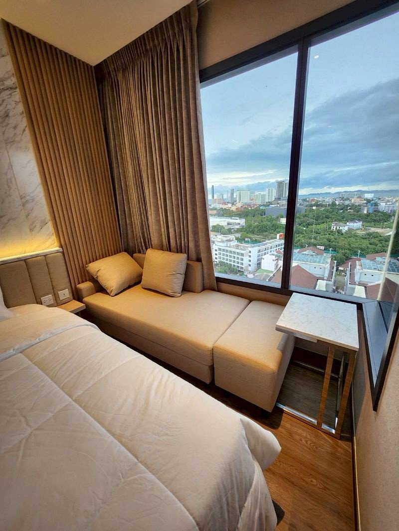 Hotel room corner with a beige sofa bed by a large window overlooking a cityscape. Fixed curtains frame the view.