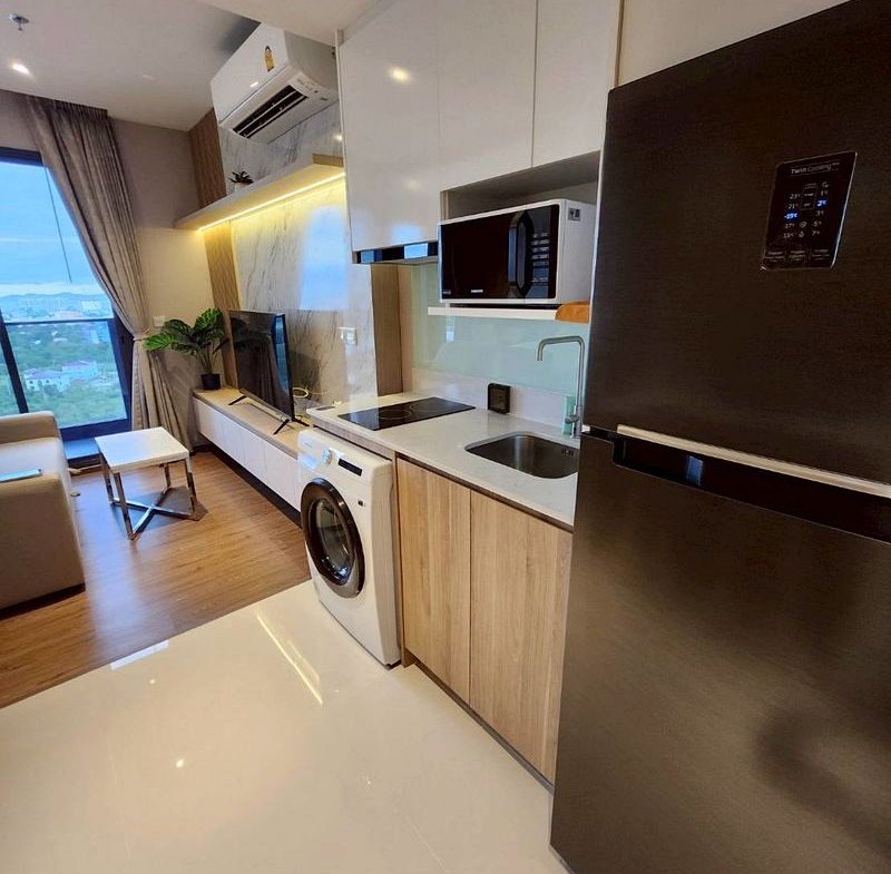 Open-plan kitchen with stainless fridge, washing machine, microwave, and a sink, leading to a living area with a TV and balcony view.