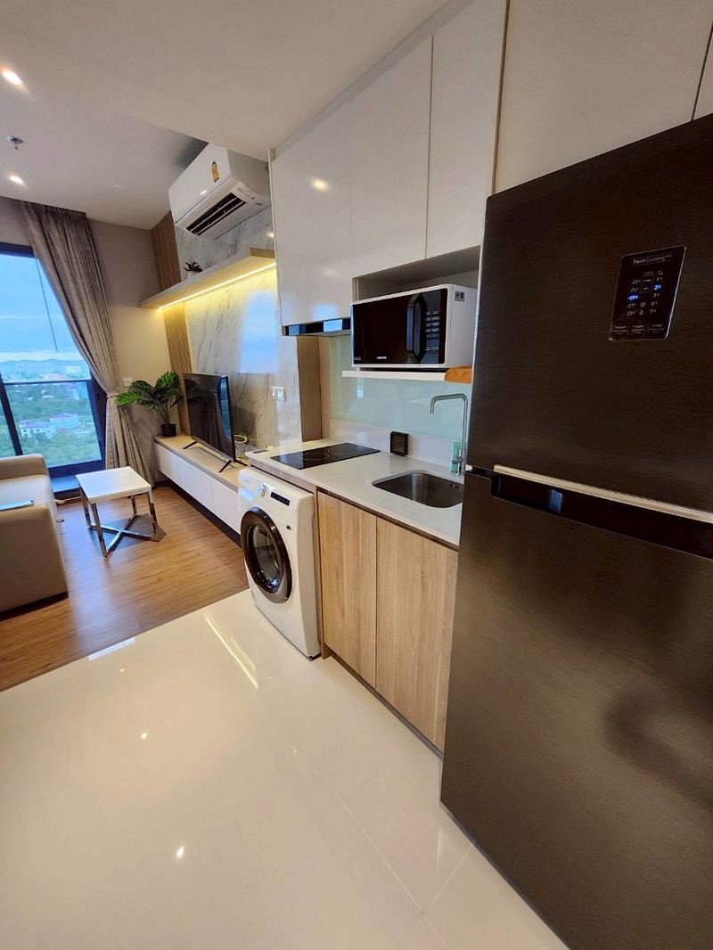 Open-plan kitchen with stainless fridge, washing machine, microwave, and a sink, leading to a living area with a TV and balcony view.
